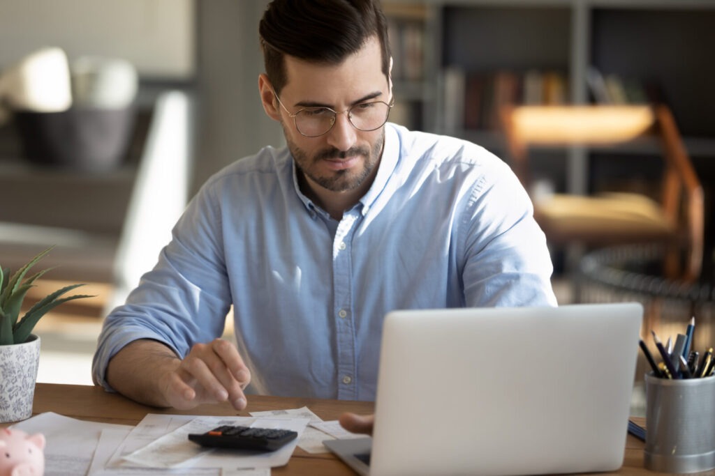 focusedyoungcaucasianmanlookatlaptopscreencalculateexpenses