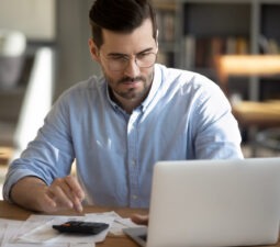 focusedyoungcaucasianmanlookatlaptopscreencalculateexpenses